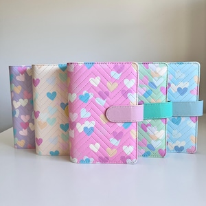 May include: Five heart-patterned planner covers in pastel shades. The covers have a quilted chevron design with heart accents. Each cover has a matching closure strap. The covers are suitable for A5 or similar sized inserts.