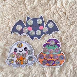 May include: Three Halloween-themed stickers: a bat, a ghost, and a frog in a pumpkin. Each sticker has holographic circles. The bat is dark blue with pink accents. The ghost is white with an orange belly. The frog is green with a purple hat.