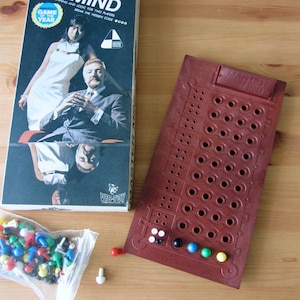 May include: A vintage Master Mind board game. The box shows a man and woman in formal wear. The reddish-brown game board has many holes. A bag of coloured pegs and loose pegs are also visible. The game's title is displayed on the box.