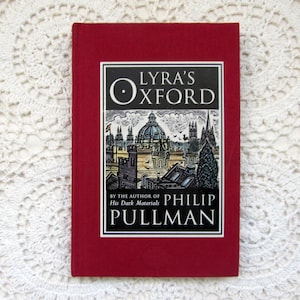 May include: A red hardback book, "Lyra's Oxford" by Philip Pullman. The cover features a black and white illustration of a city with a domed building. The title is in a black box. The book is on a white lace surface.