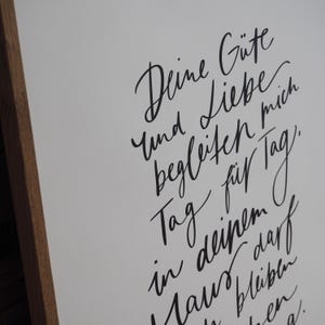 May include: A framed white print with black calligraphy. The text reads "Deine G&uuml;te und Liebe begleiten mich Tag f&uuml;r Tag in deinem Haus bleiben". The frame is made of brown wood.