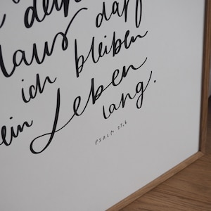 May include: A framed print featuring black calligraphy on a white backdrop. The German text includes "ich Leben lang" and the reference "Psalm 23,6". The frame is constructed from light brown wood, adding a natural touch to the artwork.