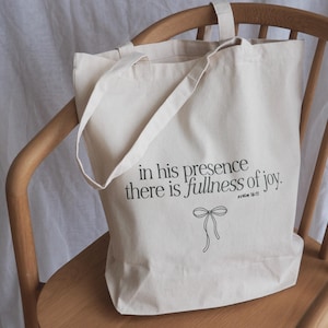 Op de afbeelding: Crèmekleurige draagtas met de tekst "in his presence there is fullness of joy" en een strikillustratie. De tas heeft twee handvatten en staat op een houten stoel.