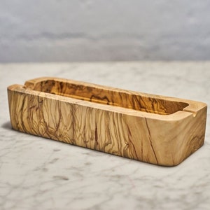 Könnte beinhalten: Eine rechteckige Holzschale mit natürlicher Holzmaserung. Die Schale ist aus Olivenholz gefertigt und hat eine glatte, polierte Oberfläche.