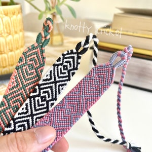 May include: Three handmade friendship bracelets in shades of pink, green, black, and white. The bracelets feature geometric patterns and braided ends. The text "knotty cricket" is visible in the background.