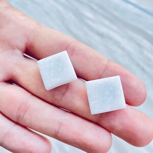 May include: A pair of square, pale blue stud earrings. Each earring is a square shape with a slightly textured surface. The earrings are displayed on a light background.