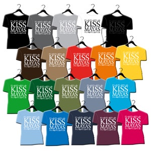 May include: Twenty-four t-shirts hanging on hangers, each with the text "KISS MAYAS" printed in white on a different colored shirt. The shirts are in various colors, including red, blue, green, yellow, orange, brown, and black.
