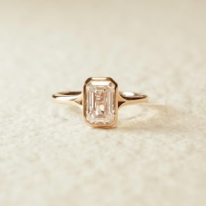 Op de afbeelding: Een gouden ring met een smaragdgeslepen diamant. De diamant is gezet in een bezel-zetting en de band is van eenvoudig, gepolijst goud. De ring wordt gepresenteerd tegen een neutrale achtergrond, waardoor het ontwerp en de schittering worden benadrukt.