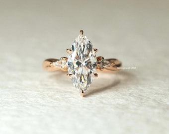 May include: A rose gold engagement ring with a prominent marquise-cut diamond. The diamond is secured by prongs and has smaller stones on either side. The band has a twisted design. The text "gemsjewelryRM" is visible. The ring is a symbol of commitment.