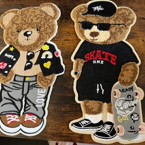 May include: Two brown teddy bear patches with embroidered details. One bear is wearing a black leather jacket with colourful patches, grey jeans, and red and white trainers. The other bear is wearing a black t-shirt with "SKATE HME" in red lettering, black shorts, white socks, and black trainers. The second bear is holding a skateboard with a skull graphic and the words "READY TO ROLL" and "GO!"
