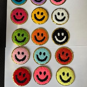 May include: A set of 15 colourful smiley face patches with a gold glitter outline. The patches are made of fuzzy fabric and have black embroidered smiley faces.