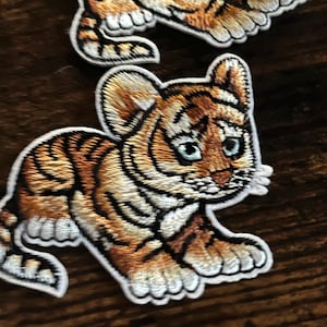 May include: Two embroidered iron-on patches of cute baby tigers. The tigers are orange and black with white stripes and white accents. The tigers are in a playful pose with their paws outstretched.