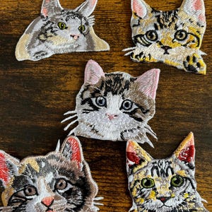May include: Five embroidered cat patches in various colours and patterns. The patches feature detailed facial features, including whiskers and eyes. The cats are in shades of grey, brown, yellow, and white, with pink accents on the ears and noses. The patches are on a brown wooden surface.