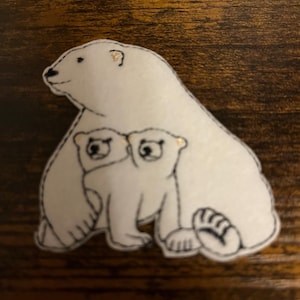 May include: White felt applique of a polar bear with two cubs. The bear is standing with its cubs nestled in its arms. The applique is outlined in black thread.