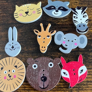 May include: A selection of wooden animal-shaped buttons. The buttons depict a cat, raccoon, zebra, rabbit, giraffe, elephant, lion, bear, and fox. Each button is painted with bright colours and detailed features, perfect for craft projects.