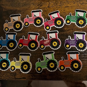 May include: Twelve colourful embroidered iron-on patches of tractors. The tractors are in various colours including red, blue, green, yellow, purple, and pink.