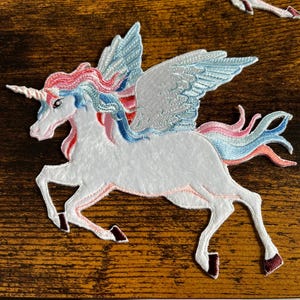 May include: A white unicorn with blue and pink wings and a pink and blue mane. The unicorn is running and has a gold horn. The unicorn is embroidered on a white fabric.