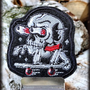 Può includere: Una toppa ricamata nera con uno scheletro argentato che fa skateboard. Lo scheletro indossa una bandana rossa e un casco argentato. La toppa ha uno sfondo nero con stelle bianche.