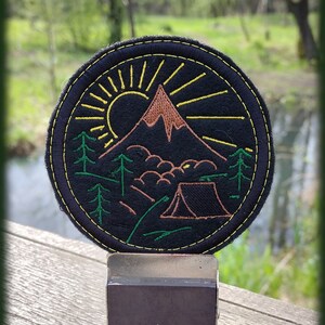 Può includere: Una toppa in feltro nero con ricamo verde e giallo che rappresenta una scena di montagna con un sole, alberi e una tenda. La toppa è fissata a un supporto magnetico argento e nero.