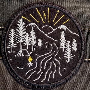 Op de afbeelding: Een zwarte geborduurde patch met een wit ontwerp van een bergketen, bomen, een rivier, een kampvuur en een tent. De zon wordt weergegeven door gele stralen.