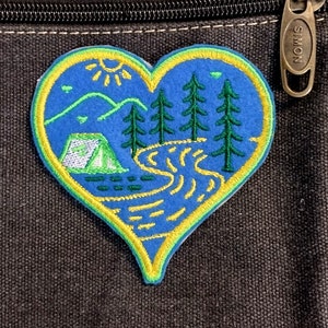 Könnte beinhalten: Herzförmiger Aufnäher mit einer Campingszene. Der Aufnäher hat einen blauen Hintergrund mit einem grünen Zelt, Bäumen und einem gelben Fluss. Das Design ist gelb und grün umrandet, mit einer Sonne und Bergen im Hintergrund.