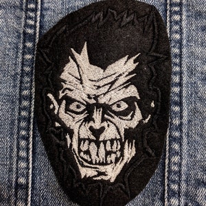 Puede incluir: Parche bordado negro con la imagen de una cara de monstruo con dientes afilados y una expresión amenazante. El parche tiene forma ovalada con un fondo negro y detalles de hilo blanco. El parche está cosido a una chaqueta vaquera.