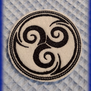 Könnte beinhalten: Ein weißer Filzkreis mit einem schwarzen gestickten Dreifachspiralenmuster. Das Muster ist ein keltischer Knoten mit drei ineinandergreifenden Spiralen.