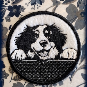 Może przedstawiać: Naszywka haftowana w kolorze czarnym i białym przedstawiająca psa zaglądającego zza powierzchni. Pies ma białą twarz i czarne futro. Naszywka jest okrągła i ma czarną obwódkę.