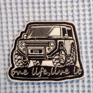 Op de afbeelding: Zwart-witte geborduurde patch van een jeep met de tekst "Live Life, Live It" eronder.