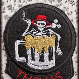 Op de afbeelding: Zwart-witte geborduurde patch van een skelet dat een rode hoed draagt en een bierpul houdt. De pul is gevuld met een gouden vloeistof en de tekst "THOMAS" is in rood onder de pul geborduurd.