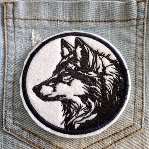Puede incluir: Un parche bordado blanco y negro de la cabeza de un lobo de perfil, cosido en un bolsillo de mezclilla azul.