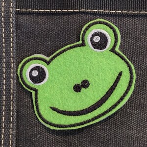 Könnte beinhalten: Grünfilz-Frosch-Applikation mit schwarzen gestickten Augen und einem Lächeln. Der Frosch ist auf einen schwarzen Stoffgrund genäht.