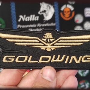 Peut inclure: Écusson brodé noir et or avec le mot "GOLDWING" et un emblème ailé. L'écusson est rectangulaire avec une bordure dorée et est tenu dans une main.