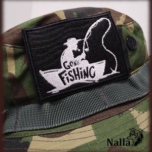 Può includere: Un cappello boonie mimetico con una toppa nera che dice "Gone Fishing" in testo bianco. La toppa presenta una silhouette di una persona che pesca da una barca.