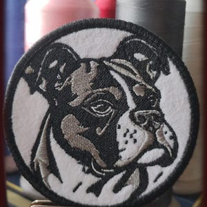 Può includere: Toppa ricamata con un volto di bulldog nero e grigio su sfondo bianco circolare. La toppa ha un bordo nero. L'immagine è ambientata su uno sfondo di rocchetti di filo colorati.