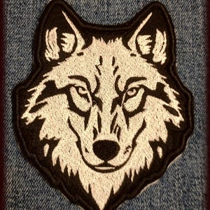 Könnte beinhalten: Gestickter Aufnäher mit einem Wolfskopf-Design. Der Wolf ist in weißem Faden mit schwarzer Umrandung dargestellt, wodurch die detaillierten Merkmale hervorgehoben werden. Der Aufnäher befindet sich auf einem Jeansstoff-Hintergrund.