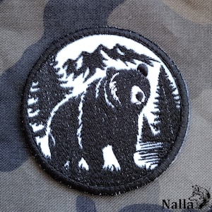 Può includere: Toppa ricamata in bianco e nero di un orso in piedi di fronte a una catena montuosa con alberi su entrambi i lati. La toppa è circolare e ha un bordo nero.