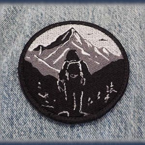 Peut inclure: Écusson rond noir et blanc représentant une montagne et un randonneur avec un sac à dos. Le motif est placé sur un fond en denim bleu clair. L'écusson est probablement à repasser ou à coudre.