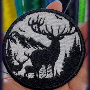 Puede incluir: Un parche bordado en blanco y negro que presenta una silueta de un ciervo con grandes cuernos y un ciervo más pequeño en primer plano. Los ciervos están de pie en un entorno de bosque con árboles en el fondo. El parche es circular y tiene un borde negro.