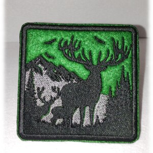 May include: A green embroidered patch with a black silhouette of a deer with large antlers standing in front of a mountain range. The deer is facing right and the mountain range is in the background. The patch has a black border.