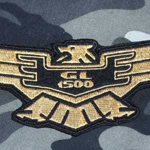 Könnte beinhalten: Gestickter Aufnäher mit einem gold-schwarzen Adlerdesign und dem Text "GL 1500". Der Aufnäher befindet sich auf einem Tarnmuster. Der Adler hat ausgebreitete Flügel und einen stilisierten Kopf.