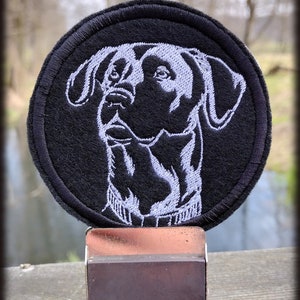 Op de afbeelding: Zwart-witte geborduurde patch van een hondenkop op een zwarte ronde achtergrond. De patch is bevestigd aan een kleine zilveren metalen standaard.