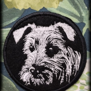 Op de afbeelding: Een geborduurde zwarte en witte patch met het gezicht van een hond. De hond heeft witte vacht en een zwarte neus. De patch is rond en heeft een zwarte rand.