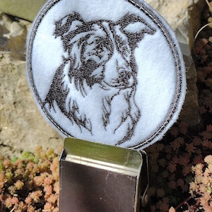 Puede incluir: Parche de fieltro blanco con una imagen bordada en negro de la cabeza de un perro Border Collie. El parche está unido a un clip de metal marrón.