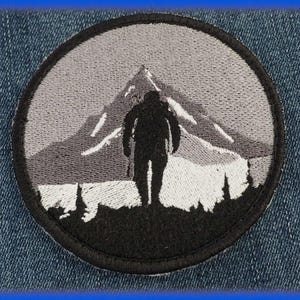 Peut inclure: Écusson brodé représentant une silhouette noire d'une personne marchant vers une chaîne de montagnes. Le motif est en nuances de gris et de noir, dans un cadre circulaire. L'écusson est sur un fond en jean.