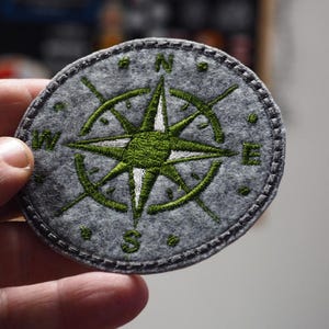 Op de afbeelding: Ronde grijze vilten patch met een groen en wit geborduurd kompasontwerp. Het kompasroos heeft richtingaanwijzers en een centrale ster. De patch heeft een gestikte rand en is een decoratief item.