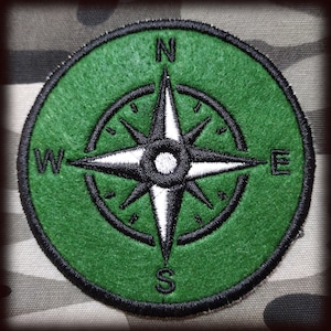 Könnte beinhalten: Ein grüner gestickter Kompass-Patch mit einer schwarzen Kompassrose und weißen Spitzen. Die Kompasspunkte sind mit N, S, E und W beschriftet.
