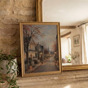 May include: A framed rectangular wall art print depicting a quiet autumn street scene at dusk. The image features houses with glowing windows, trees with orange foliage, street lamps, and jack-o'-lanterns lining a path in a gold frame.
