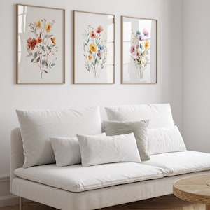 May include: Three framed watercolour paintings of colourful flower bouquets with green leaves. The paintings are hung on a white wall above a white sofa with white cushions.