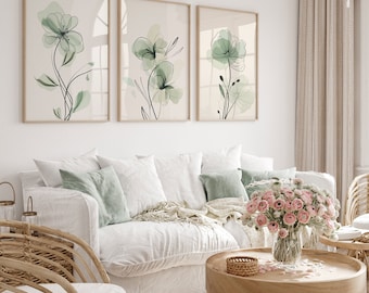 May include: Three framed watercolor paintings of green flowers with thin black stems on a white wall above a white couch with pillows and a throw blanket. A bouquet of pink flowers in a glass vase sits on a wooden coffee table.